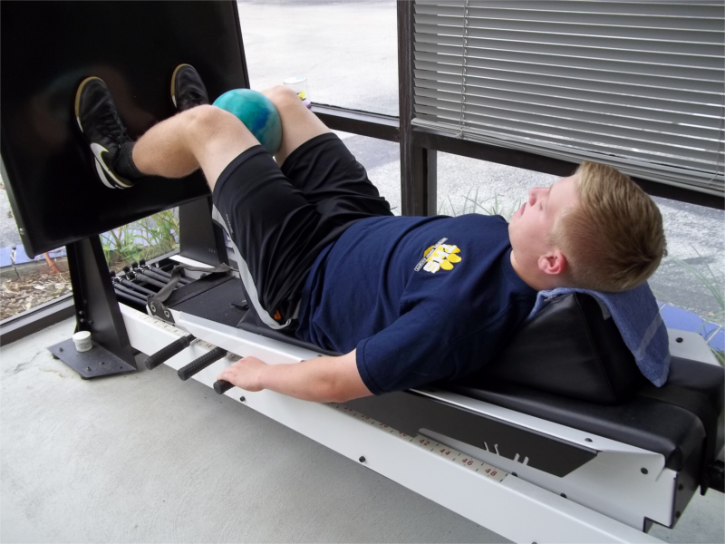 Young Man with Ball Between Knees, using Shuttle MVP Machine. Lying Down while Pushing in Squat like position.